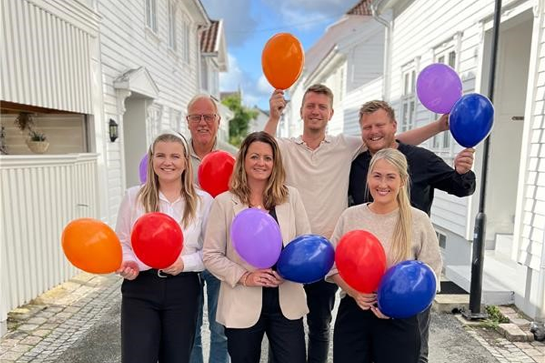 Seks ansatte i Sparebanken Norge står utenfor hvitmalte trehus med fargerike ballonger