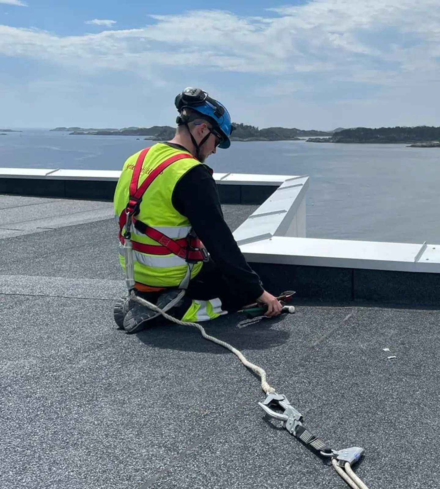 Takarbeider sikret med sele sitter på et flatt tak med utsikt over sjø, strand og skog
