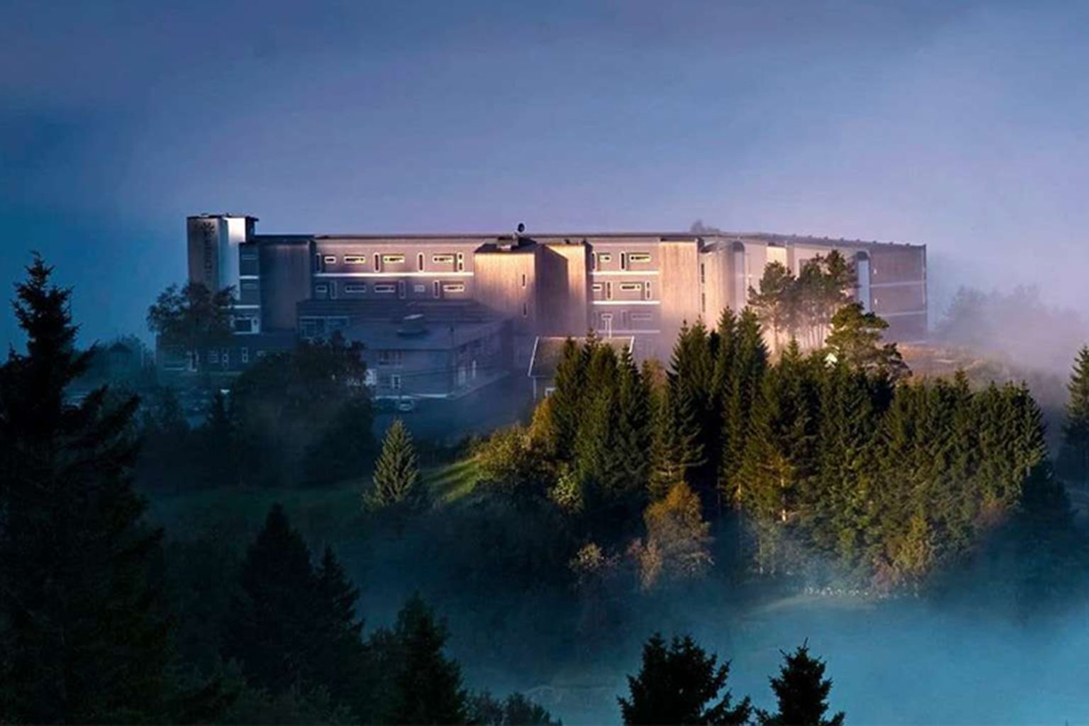 Utsikten Hotell sett utenfra i kveldslys, med skogen og tåke rundt bygningen