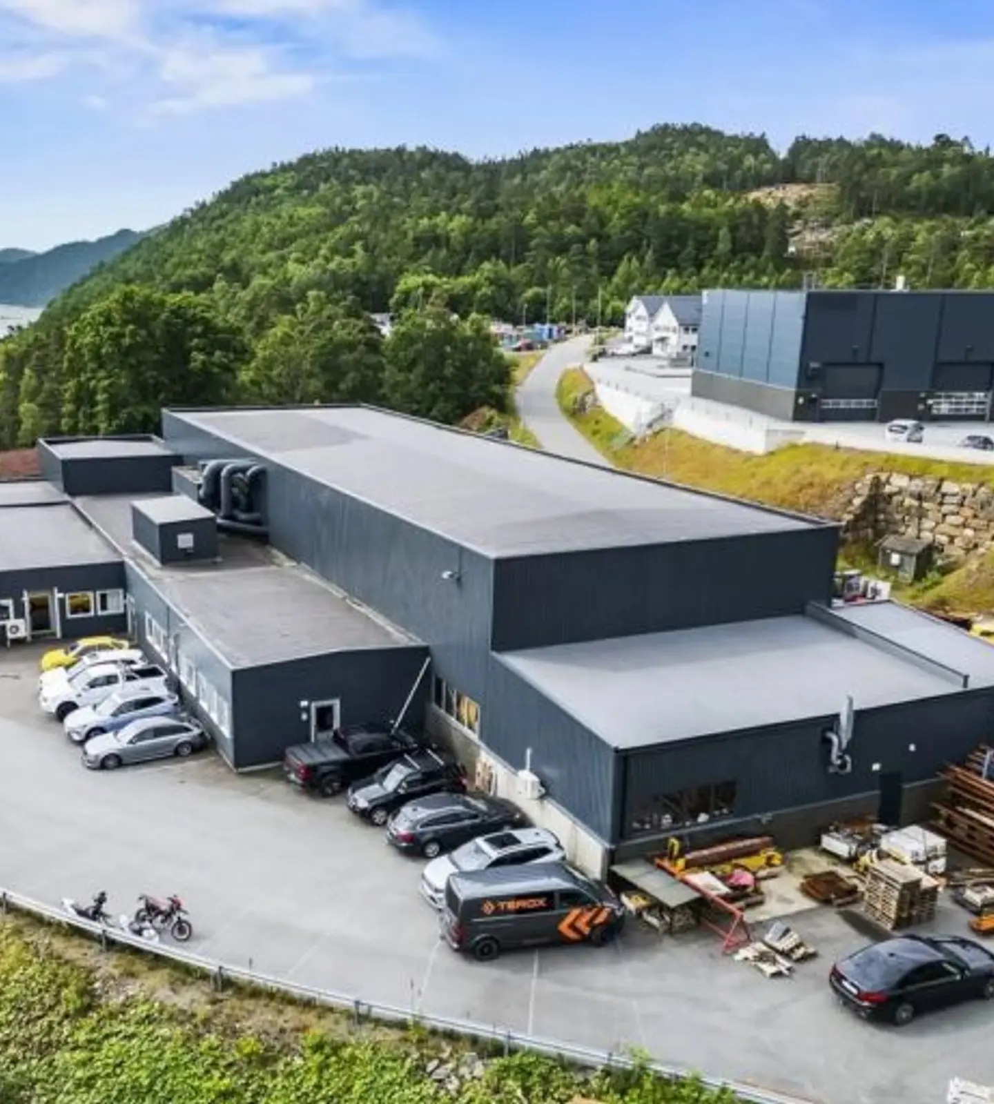 Luftfoto av Terox sitt industribygg i Svegeskogen, med parkerte biler, en anleggsmaskin og utsikt over fjorden