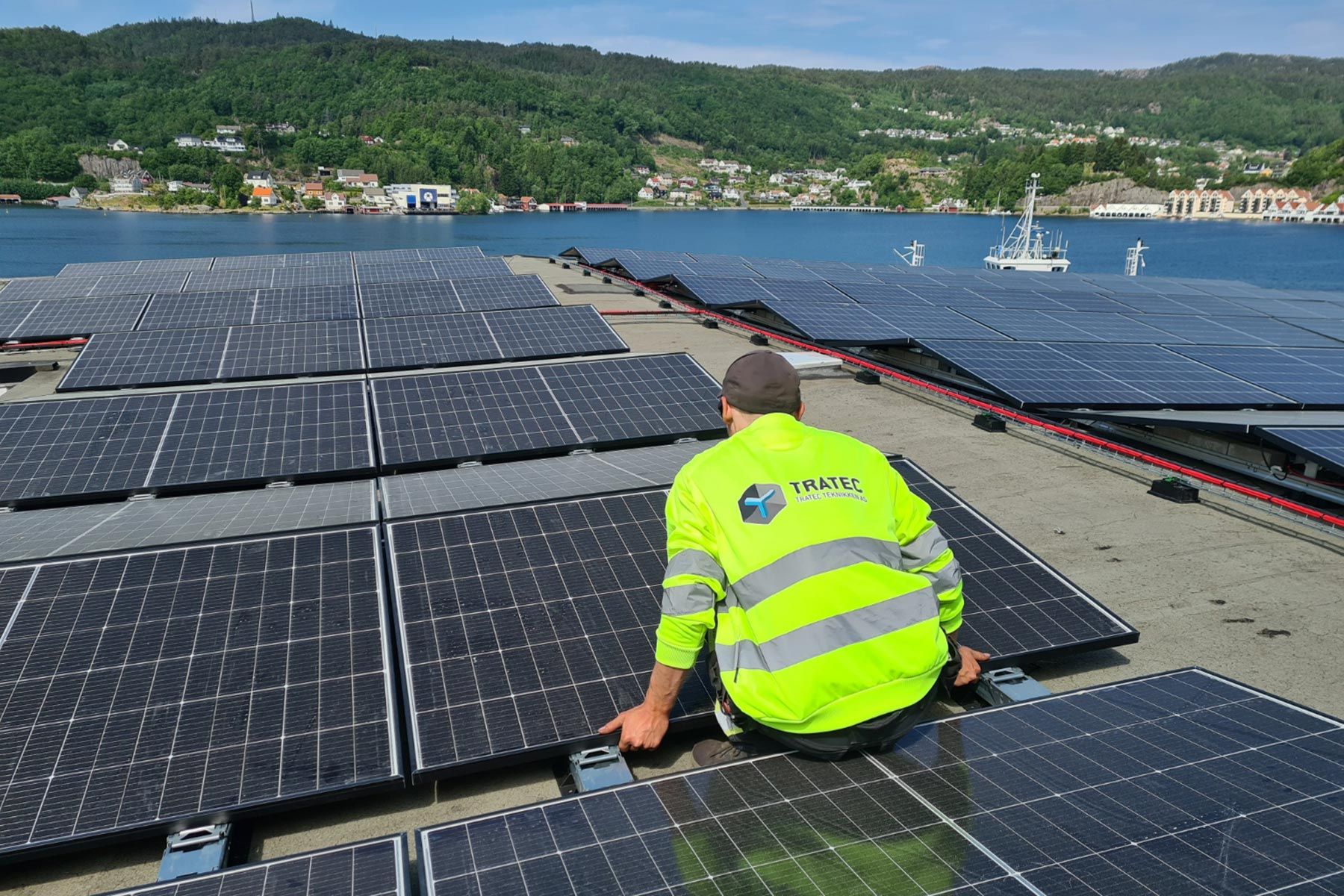 En ansatt i TRATEC-jakke monterer solcellepaneler på et flytende anlegg, med en fjordby i bakgrunnen.