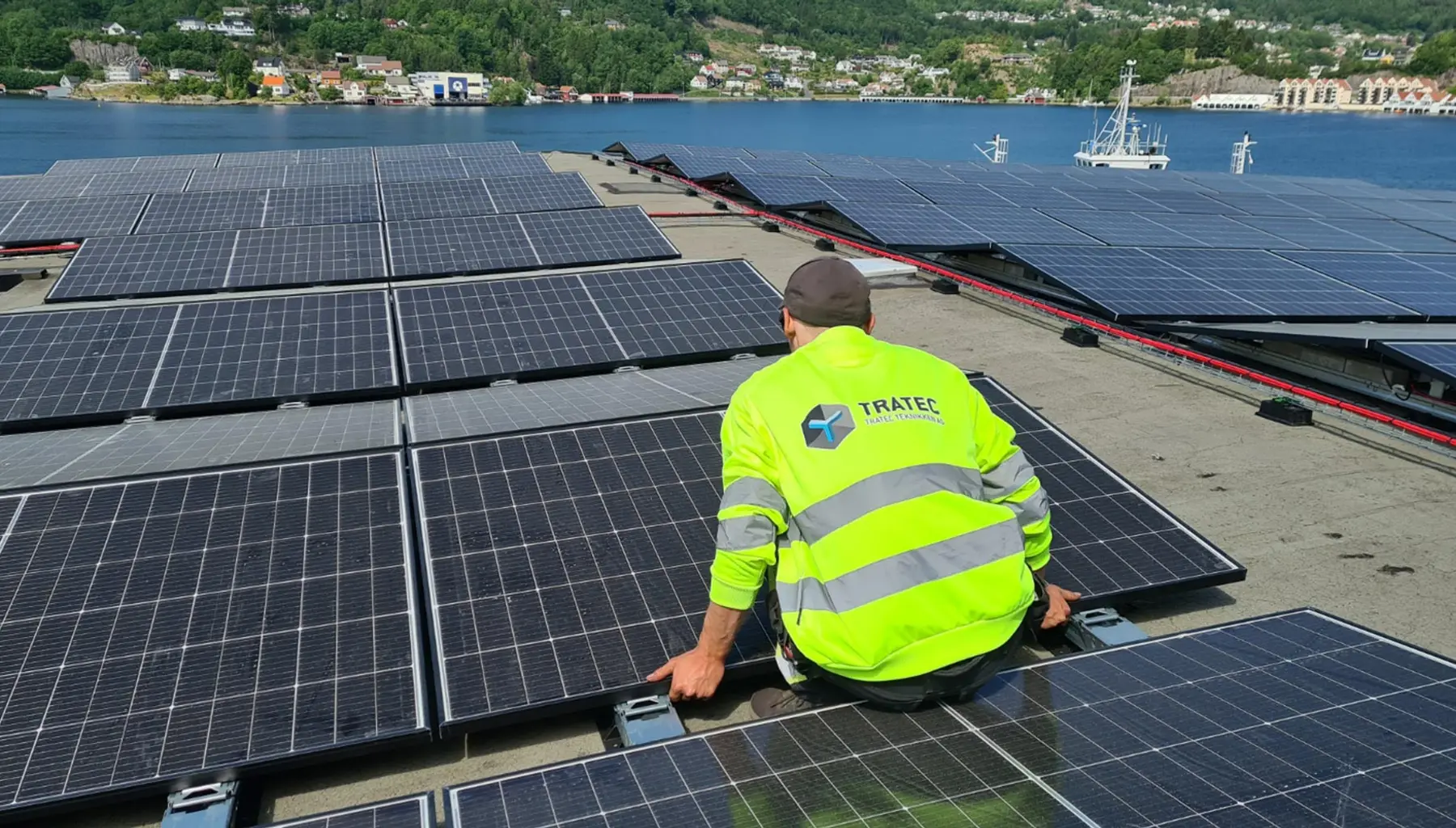 En ansatt i TRATEC-jakke monterer solcellepaneler på et flytende anlegg, med en fjordby i bakgrunnen.