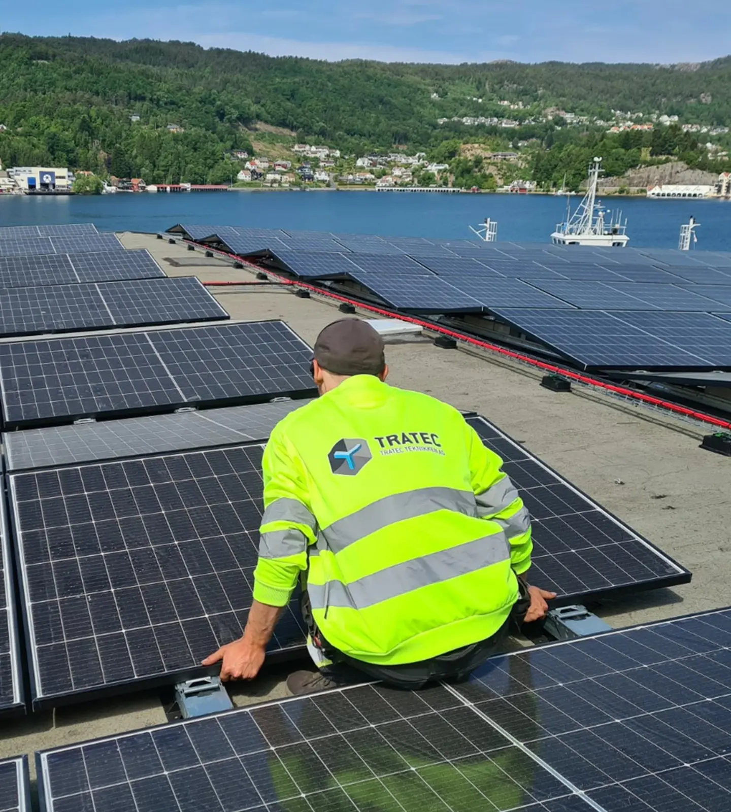 En ansatt i TRATEC-jakke monterer solcellepaneler på et flytende anlegg, med en fjordby i bakgrunnen.