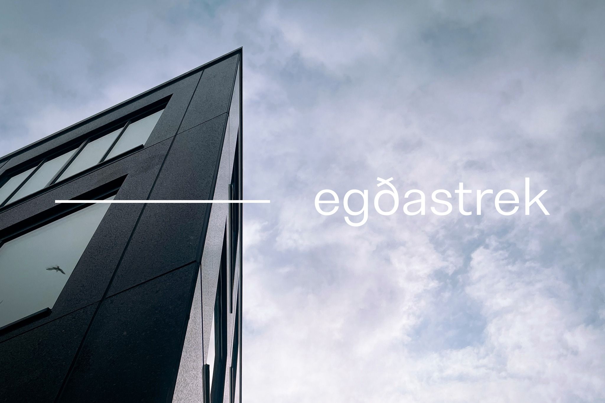 Hjørnet av en mørk, moderne bygning sett nedenfra mot skyet himmel, med egðastreks logo