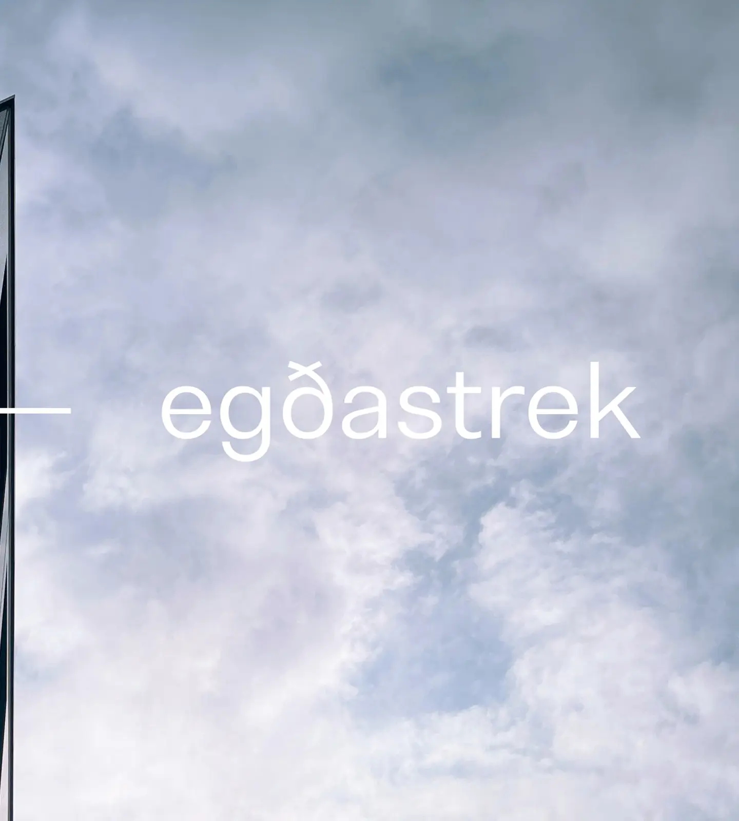 Hjørnet av en mørk, moderne bygning sett nedenfra mot skyet himmel, med egðastreks logo