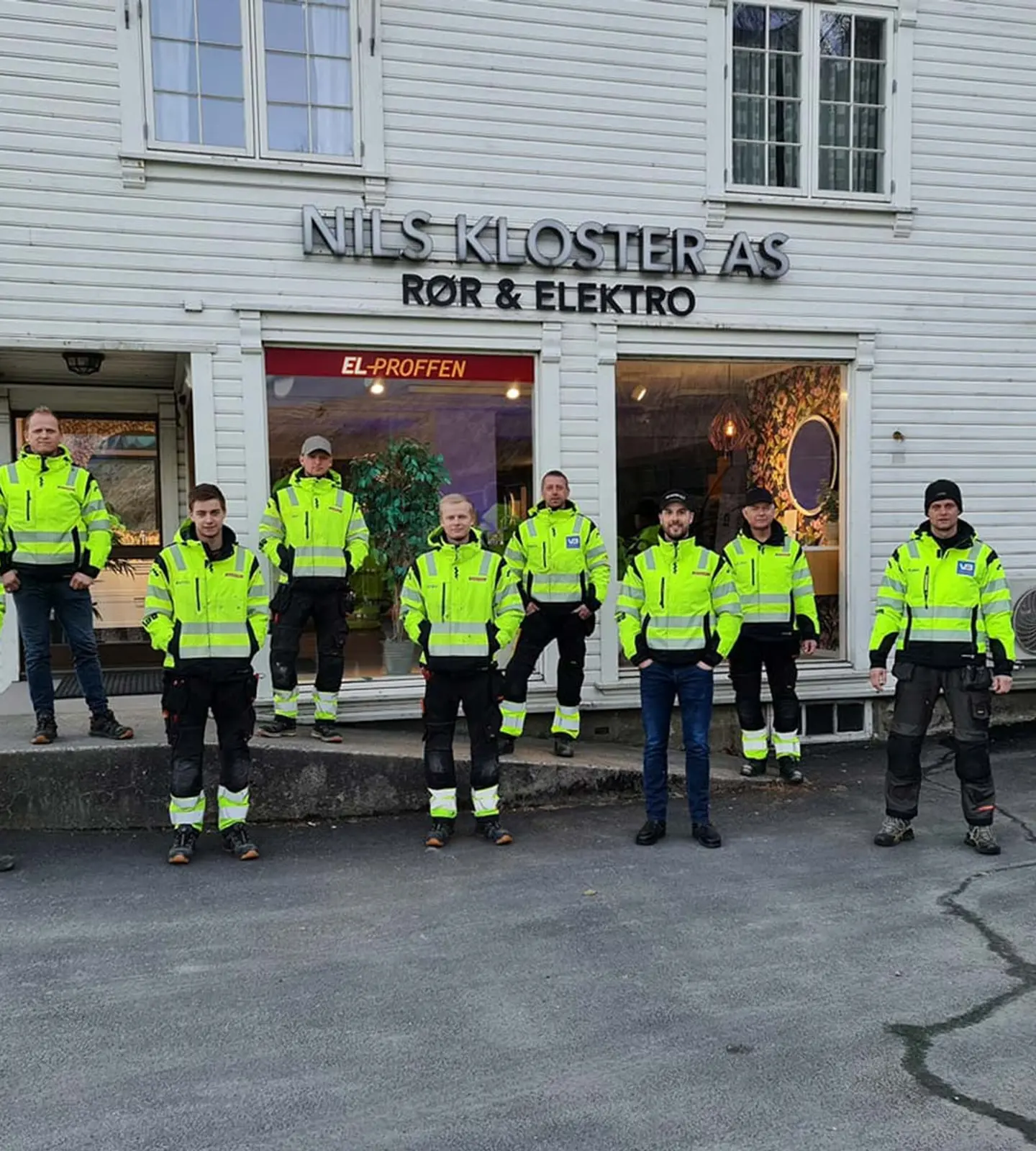 Ni ansatte i gule varseljakker fra Nils Kloster AS står samlet foran bedriftens hvite bygning med skilt for rør og elektro i Kvinesdal.