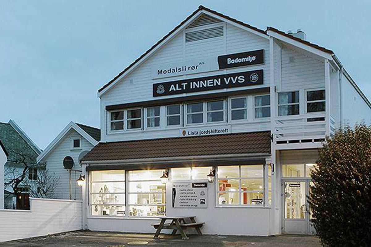 Bademiljø Modalsli Rørs hvite bygning i Flekkefjord sentrum, med skilt «Alt innen VVS» og opplyste utstillingsvinduer i kveldsmørket