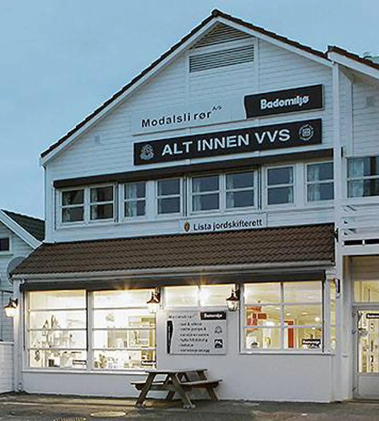 Bademiljø Modalsli Rørs hvite bygning i Flekkefjord sentrum, med skilt «Alt innen VVS» og opplyste utstillingsvinduer i kveldsmørket